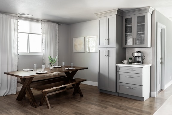 A modern kitchen and dining area featuring light gray cabinets with glass-paneled doors, a dark wooden dining table with matching benches, and decoratively arranged dishes and glasses. A coffee machine sits on a white countertop, and a potted plant is placed in the center of the dining table. Soft, natural light filters through white draped curtains covering a window.