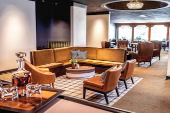 A stylish lounge area with a large U-shaped tan couch and a round coffee table in the center. There are several modern chairs around the table, and large windows bring in natural light. A decorative chandelier hangs above, enhancing the sophisticated ambiance. A tray with glasses and a decanter is prominently displayed in the foreground.