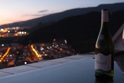 A bottle of Viña Sol wine is placed on a table with a stunning cityscape in the background during twilight. The city lights create a bokeh effect, with a mountain range silhouetted against the fading sunset.