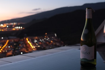A bottle of Vi&ntilde;a Sol wine is placed on a table with a stunning cityscape in the background during twilight. The city lights create a bokeh effect, with a mountain range silhouetted against the fading sunset.
