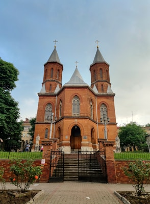 A red brick church with two tall towers topped with crosses is surrounded by a black iron fence. Stairs lead to the entrance, flanked by statues. The sky is mostly clear, and there are rose bushes in front of the church.