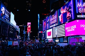 A bustling city street scene illuminated by numerous bright, colorful billboards and advertisements. The street is filled with people, creating a lively and energetic atmosphere. The advertisements range from entertainment shows to retail promotions, contributing to the vibrant urban setting.
