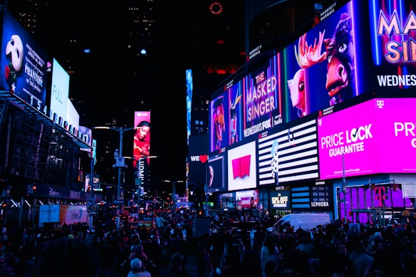 A bustling city street scene illuminated by numerous bright, colorful billboards and advertisements. The street is filled with people, creating a lively and energetic atmosphere. The advertisements range from entertainment shows to retail promotions, contributing to the vibrant urban setting.