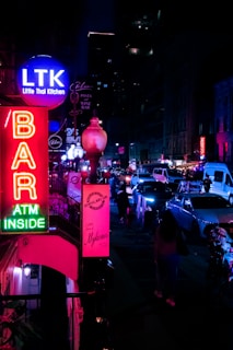 A bustling urban street scene at night with vibrant neon signs illuminating the area. The signs advertise various establishments, including a Thai kitchen and a bar. Vehicles can be seen parked and driving through the street, while some pedestrians walk by. The overall ambiance is lively, with a mix of modern city life and nightlife elements.