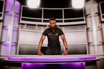 A person is standing behind a modern desk, leaning forward with both hands resting on the desk. The setting resembles a futuristic or high-tech environment, featuring sleek metallic panels and purple lighting. The person is wearing a dark polo shirt and a watch, and they have a focused expression.