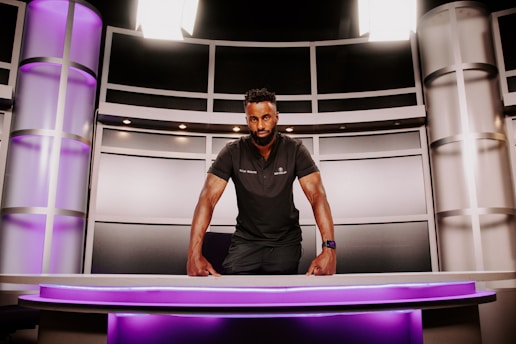 A person is standing behind a modern desk, leaning forward with both hands resting on the desk. The setting resembles a futuristic or high-tech environment, featuring sleek metallic panels and purple lighting. The person is wearing a dark polo shirt and a watch, and they have a focused expression.