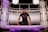 A person is standing behind a modern desk, leaning forward with both hands resting on the desk. The setting resembles a futuristic or high-tech environment, featuring sleek metallic panels and purple lighting. The person is wearing a dark polo shirt and a watch, and they have a focused expression.
