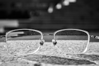 A pair of eyeglasses with rectangular frames rests on a textured stone surface. The background is blurred, giving a sense of depth, and the glasses themselves have visible tiny droplets or marks on the lenses.