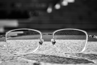 A pair of eyeglasses with rectangular frames rests on a textured stone surface. The background is blurred, giving a sense of depth, and the glasses themselves have visible tiny droplets or marks on the lenses.