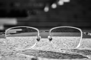 A pair of eyeglasses with rectangular frames rests on a textured stone surface. The background is blurred, giving a sense of depth, and the glasses themselves have visible tiny droplets or marks on the lenses.