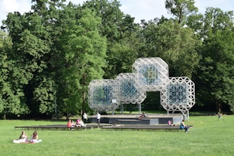 A geometric, lattice-like structure made of interconnected modules stands prominently in a park surrounded by lush green trees. Several people are sitting, lying, or walking on the grass area nearby. This open area suggests a place for leisure and outdoor activities.