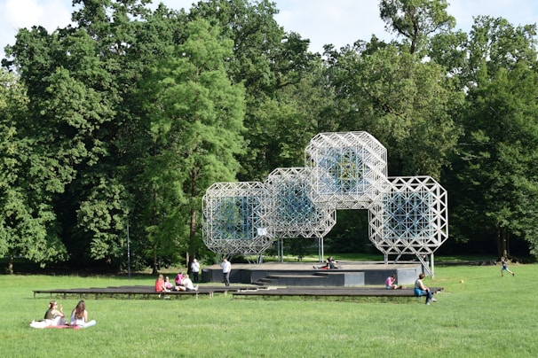 A geometric, lattice-like structure made of interconnected modules stands prominently in a park surrounded by lush green trees. Several people are sitting, lying, or walking on the grass area nearby. This open area suggests a place for leisure and outdoor activities.