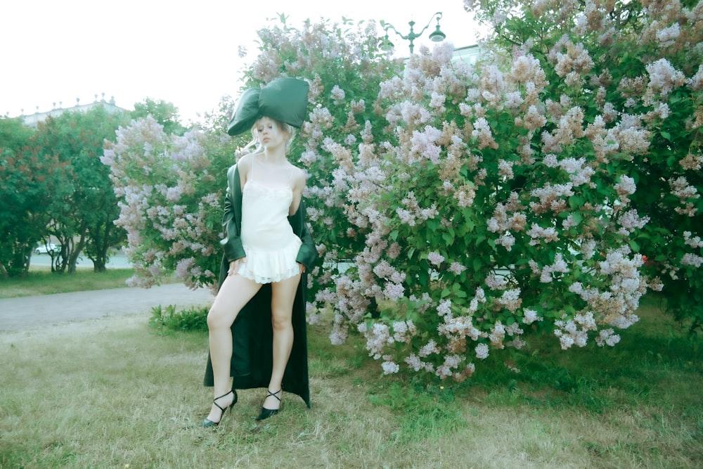 a woman in a dress and hat standing in front of a bush