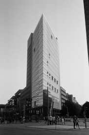 A tall, modern skyscraper with a sharp, angular design dominates the cityscape. The facade is composed of reflective glass panels that capture the light, emphasizing the building's height. Below the structure, pedestrians and cyclists move along the bustling streets, suggesting a lively urban setting. To the left, a sign for Potsdamer Platz is visible, indicating the location. The image is monochrome, adding a timeless and classic feel.