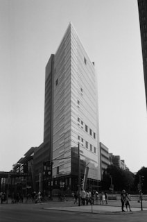 A tall, modern skyscraper with a sharp, angular design dominates the cityscape. The facade is composed of reflective glass panels that capture the light, emphasizing the building's height. Below the structure, pedestrians and cyclists move along the bustling streets, suggesting a lively urban setting. To the left, a sign for Potsdamer Platz is visible, indicating the location. The image is monochrome, adding a timeless and classic feel.