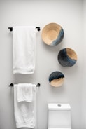 A bathroom wall with three decorative woven baskets featuring spiral patterns in neutral and dark tones. Two white towels are neatly hanging on black metal towel racks mounted on the same wall. Below the towel racks, there is a portion of a white toilet visible.