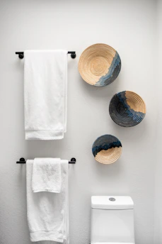 A bathroom wall with three decorative woven baskets featuring spiral patterns in neutral and dark tones. Two white towels are neatly hanging on black metal towel racks mounted on the same wall. Below the towel racks, there is a portion of a white toilet visible.
