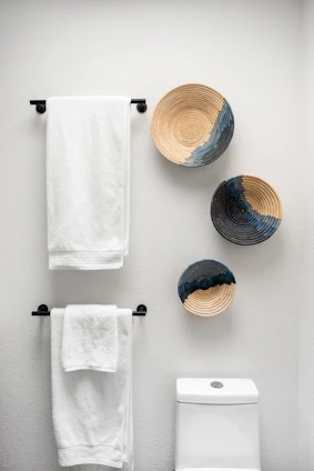 A rattan wall-mounted storage basket filled with rolled towels and small toiletries in a bright bathroom.