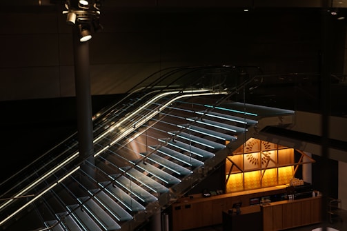 A sleek, modern staircase with glass side panels and illuminated steps ascends within a dimly lit interior space. Overhead spotlights enhance the mood with directed lighting. Below the stairs, the warmly lit counter area features geometric wooden paneling and decorative designs.