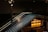 A sleek, modern staircase with glass side panels and illuminated steps ascends within a dimly lit interior space. Overhead spotlights enhance the mood with directed lighting. Below the stairs, the warmly lit counter area features geometric wooden paneling and decorative designs.