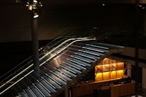 A sleek, modern staircase with glass side panels and illuminated steps ascends within a dimly lit interior space. Overhead spotlights enhance the mood with directed lighting. Below the stairs, the warmly lit counter area features geometric wooden paneling and decorative designs.