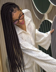 A person with long braided hair wears glasses and a textured, white garment. They are smiling and appear to be interacting with large, vertically-aligned circular panels in shades of green and gray. The setting suggests a modern, artistic or design-focused environment.