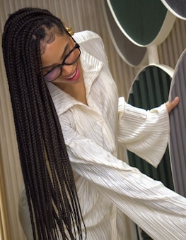 A person with long braided hair wears glasses and a textured, white garment. They are smiling and appear to be interacting with large, vertically-aligned circular panels in shades of green and gray. The setting suggests a modern, artistic or design-focused environment.