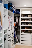 A well-organized walk-in closet featuring neatly arranged clothes on hangers, a set of drawers, and shelves filled with various pairs of shoes. Luggage bags are stored at the top, and a pillow is placed on the upper shelf.