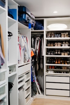 A well-organized walk-in closet featuring neatly arranged clothes on hangers, a set of drawers, and shelves filled with various pairs of shoes. Luggage bags are stored at the top, and a pillow is placed on the upper shelf.