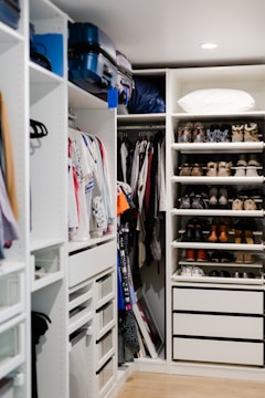 A well-organized walk-in closet featuring neatly arranged clothes on hangers, a set of drawers, and shelves filled with various pairs of shoes. Luggage bags are stored at the top, and a pillow is placed on the upper shelf.