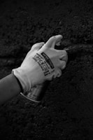 A gloved hand holding a spray paint can against a rough textured surface. The scene appears monochromatic, emphasizing the rugged texture of the wall.