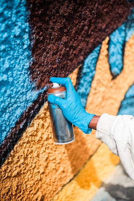 A person wearing a blue glove is holding a silver spray can and applying paint to a textured wall. The wall features vibrant colors, including blue, brown, and yellow.