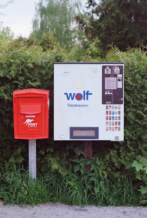 A red postbox is positioned next to a cigarette vending machine, both standing against a background of green shrubbery. The postbox is mounted on a metal pole, and the vending machine displays various colorful packs of tobacco products along with the brand label 'wolf Tabakwaren'.