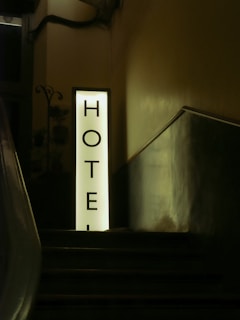 A dimly lit staircase leading up to a prominently illuminated vertical sign with the word 'HOTEL' written on it in bold letters. The surrounding walls and area are shadowed, giving a sense of mystery and intrigue.