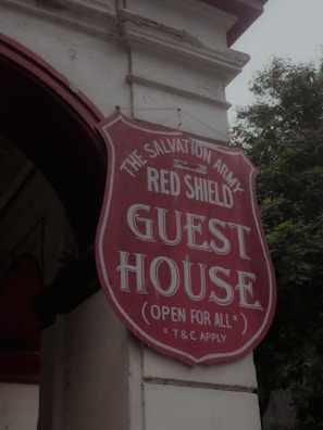 A red and white sign for The Salvation Army Red Shield Guest House is attached to the exterior of a building. The sign is shield-shaped with bold lettering and includes the phrase 'Open for All' and 'T&C Apply'. The building has a light-colored facade and an archway, with trees visible in the background.