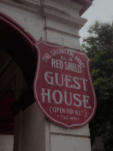A red and white sign for The Salvation Army Red Shield Guest House is attached to the exterior of a building. The sign is shield-shaped with bold lettering and includes the phrase 'Open for All' and 'T&C Apply'. The building has a light-colored facade and an archway, with trees visible in the background.
