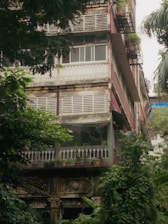 A multi-story residential building with a rustic, aged appearance surrounded by lush green foliage. The structure has detailed architectural elements such as wooden railings, decorative stonework, and potted plants throughout the balcony areas. Weathered features like peeling paint and discolored walls add to the vintage charm.