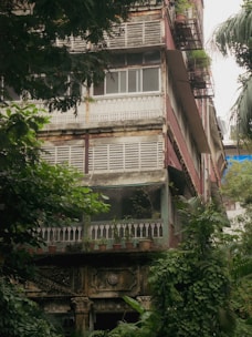 A multi-story residential building with a rustic, aged appearance surrounded by lush green foliage. The structure has detailed architectural elements such as wooden railings, decorative stonework, and potted plants throughout the balcony areas. Weathered features like peeling paint and discolored walls add to the vintage charm.