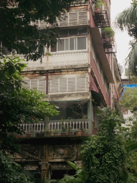 A multi-story residential building with a rustic, aged appearance surrounded by lush green foliage. The structure has detailed architectural elements such as wooden railings, decorative stonework, and potted plants throughout the balcony areas. Weathered features like peeling paint and discolored walls add to the vintage charm.