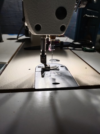 A close-up view of a sewing machine needle and presser foot, with visible stitching thread. The sewing machine is positioned on a work table, and there are subtle shadows on the surface.