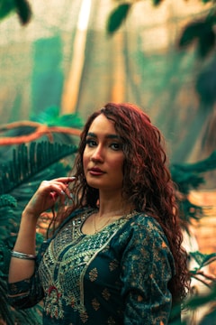 A woman with curly hair is dressed in traditional attire, standing in front of a backdrop of lush green plants. Her attire is detailed with intricate patterns, and she gazes into the camera with a confident expression.