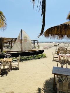 A sandy beach setting with wooden tables and chairs arranged under large palm-thatched umbrellas. A small sailboat with printed text is positioned near the seating area, surrounded by tropical plants. The beach extends towards the horizon, where sun loungers and umbrellas are visible near the ocean. Palm leaves hang overhead, adding to the tropical atmosphere.