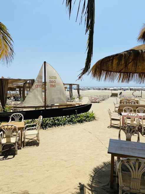 A sandy beach setting with wooden tables and chairs arranged under large palm-thatched umbrellas. A small sailboat with printed text is positioned near the seating area, surrounded by tropical plants. The beach extends towards the horizon, where sun loungers and umbrellas are visible near the ocean. Palm leaves hang overhead, adding to the tropical atmosphere.