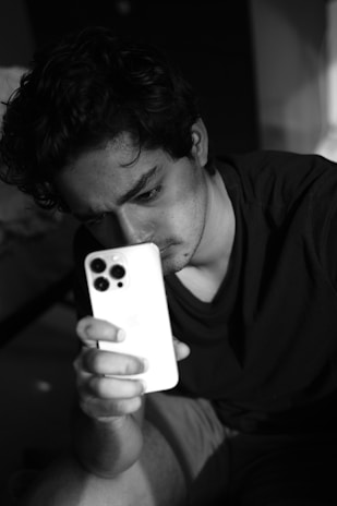 A person is sitting down, intensely looking at a smartphone in their hand. The image is in black and white, creating a dramatic contrast and mood. The lighting casts soft shadows on the person's face, emphasizing their focused expression.