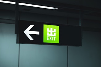 A black signboard with an illuminated green section displaying the word 'EXIT' in white text. To the left of the text, a large white arrow points to the left.