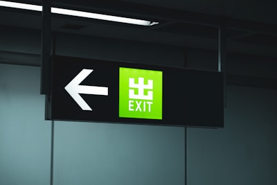A black signboard with an illuminated green section displaying the word 'EXIT' in white text. To the left of the text, a large white arrow points to the left.