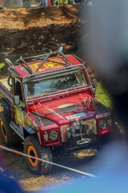 A modified red off-road vehicle, equipped with a roof rack and adorned with various sponsor logos, is participating in a rugged event. The car is navigating a muddy terrain, surrounded by blurred figures and natural elements. The wheels are specially designed for off-road driving.