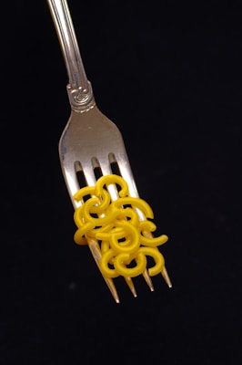 A silver fork with ornate details holds several pieces of uncooked yellow macaroni against a solid black background.