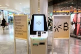 A cryptocurrency ATM is positioned in a modern shopping mall, flanked by two informational signs. The left sign lists currencies available for transaction including Bitcoin, Ethereum, Litecoin, and others, with a company logo at the bottom. The right sign invites users to exchange crypto, displaying symbols and a company logo. Shopper moving with a cart is visible in the background, along with clothing stores.