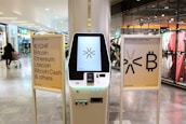 A cryptocurrency ATM is positioned in a modern shopping mall, flanked by two informational signs. The left sign lists currencies available for transaction including Bitcoin, Ethereum, Litecoin, and others, with a company logo at the bottom. The right sign invites users to exchange crypto, displaying symbols and a company logo. Shopper moving with a cart is visible in the background, along with clothing stores.